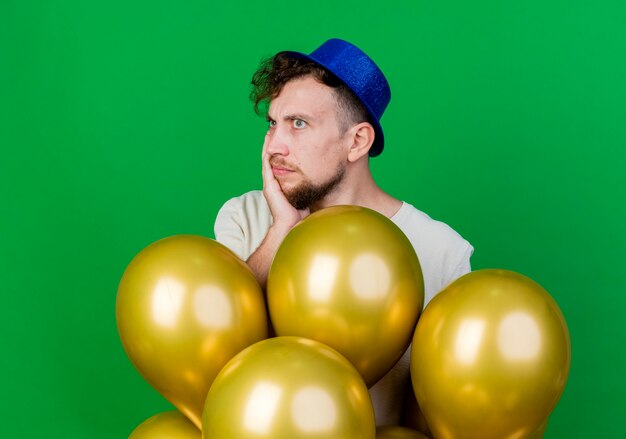 Réfléchi jeune beau mec de parti slave portant chapeau de fête debout derrière des ballons en gardant la main sur le visage à côté isolé sur fond vert