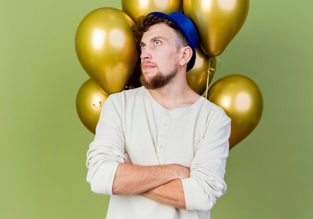 Réfléchi jeune beau mec de parti slave portant chapeau de fête debout avec une posture fermée devant des ballons à côté isolé sur mur vert olive avec espace copie