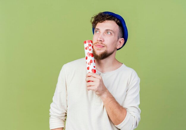 Réfléchi jeune beau mec de parti slave portant chapeau de fête touchant le visage avec canon à confettis à côté isolé sur fond vert olive avec espace de copie