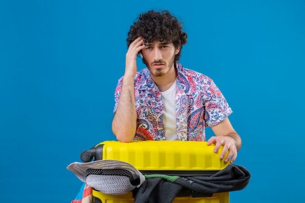 Réfléchi jeune beau voyageur bouclé homme mettant les mains sur la tête et sur une valise pleine de chiffons sur un mur bleu isolé avec copie espace