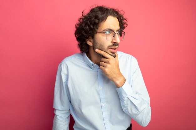 Réfléchi jeune bel homme caucasien portant des lunettes touchant le menton à côté isolé sur fond cramoisi avec espace de copie