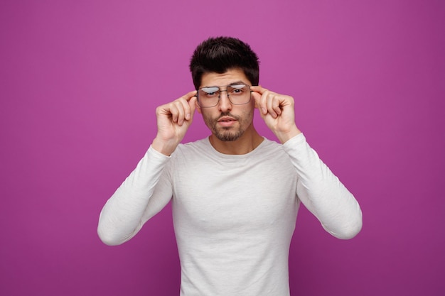 Réfléchi jeune bel homme portant et touchant des lunettes regardant la caméra sur fond violet