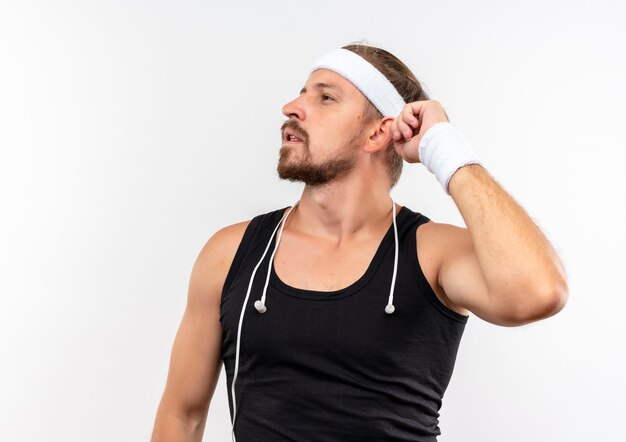 Réfléchi jeune bel homme sportif portant un bandeau et des bracelets avec des écouteurs autour de son cou en gardant la main près de la tête à côté isolé sur un espace blanc