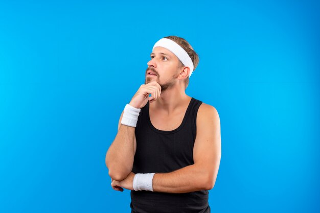 Réfléchi jeune bel homme sportif portant un bandeau et des bracelets mettant les mains sur le menton et sous le coude à côté isolé sur l'espace bleu