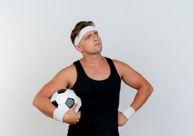 Réfléchi jeune bel homme sportif portant un bandeau et des bracelets tenant un ballon de football et mettant la main sur la taille en regardant à côté isolé sur un mur blanc