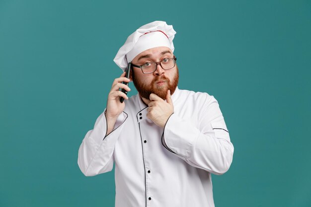 Réfléchi jeune chef masculin portant des lunettes uniformes et une casquette gardant la main sur le menton regardant la caméra parler au téléphone isolé sur fond bleu