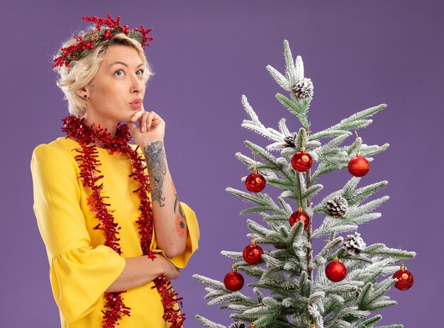 Réfléchi jeune femme blonde portant couronne de tête de Noël et guirlande de guirlandes autour du cou debout près de l'arbre de Noël décoré en gardant la main sur le menton en levant isolé sur fond violet