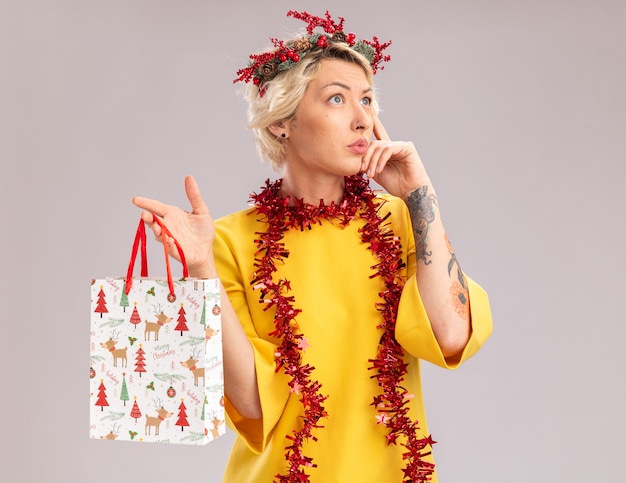 Réfléchi jeune femme blonde portant une couronne de tête de Noël et guirlande de guirlandes autour du cou tenant un sac-cadeau de Noël à côté faisant penser geste isolé sur fond blanc