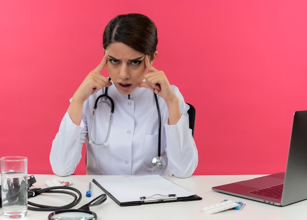 Réfléchi jeune femme médecin portant une robe médicale et un stéthoscope assis au bureau avec des outils médicaux et un ordinateur portable mettant les doigts sur les temples isolés sur le mur rose