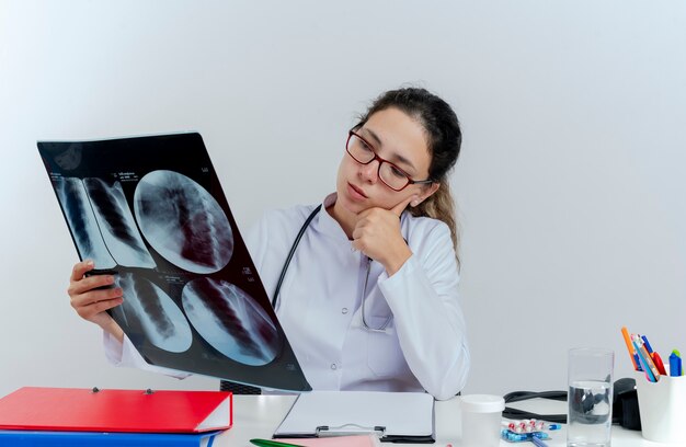 Réfléchi jeune femme médecin portant une robe médicale et un stéthoscope et des lunettes assis au bureau avec des outils médicaux tenant à la recherche de tir aux rayons x mettant la main sur le menton isolé