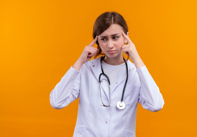 Réfléchi jeune femme médecin portant une robe médicale et un stéthoscope mettant les doigts sur les tempes sur un mur orange isolé avec copie espace