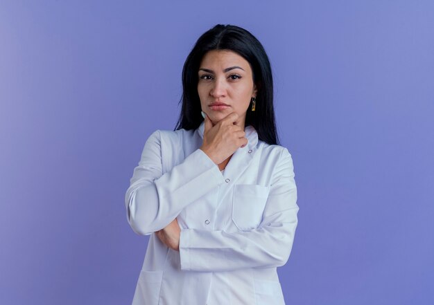 Réfléchi jeune femme médecin portant une robe médicale touchant le menton isolé sur un mur violet avec copie espace