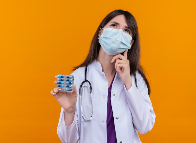 Réfléchi jeune femme médecin en robe médicale avec stéthoscope porte un masque médical jetable détient la médecine sur fond orange isolé avec copie espace