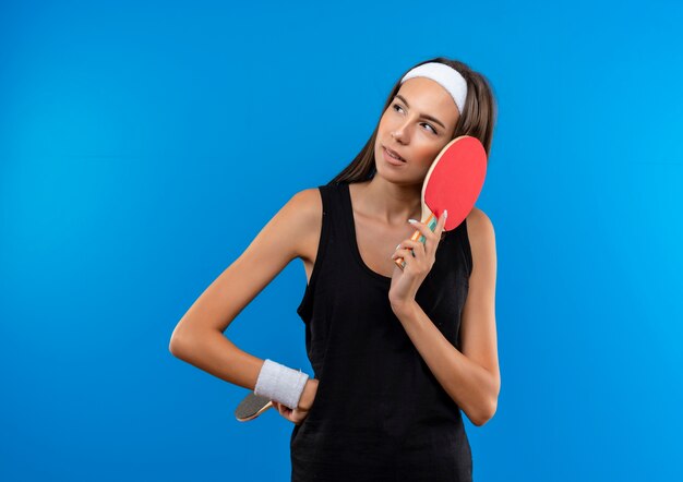 Réfléchi jeune fille assez sportive portant bandeau et bracelet tenant des raquettes de ping-pong et mettant la main sur la taille à côté isolé sur l'espace bleu