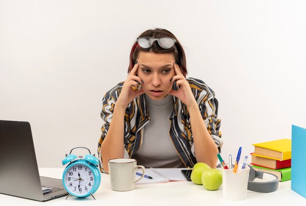 Réfléchi jeune fille étudiante portant des lunettes sur la tête assis au bureau avec des outils universitaires mettant les doigts sur les tempes à côté isolé sur mur blanc