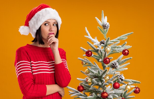 Réfléchi jeune fille portant un bonnet de Noel debout près de l'arbre de Noël décoré en gardant la main sur le menton à côté isolé sur mur orange