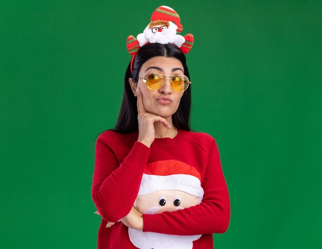 Réfléchi jeune fille de race blanche portant bandeau de père Noël et pull avec des lunettes à côté en gardant la main sur le menton à côté isolé sur fond vert avec espace de copie