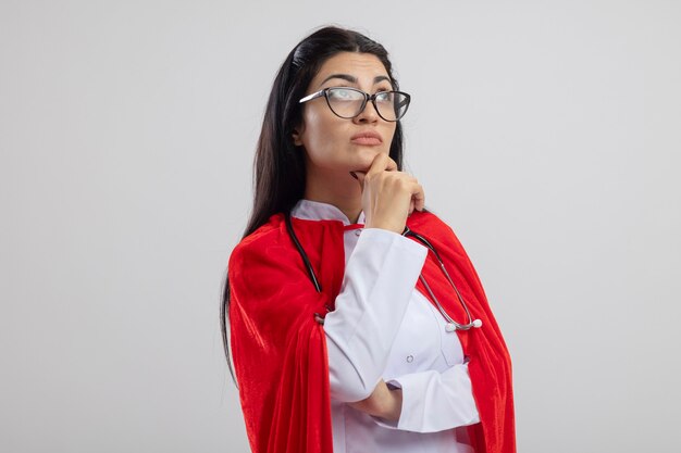 Réfléchi jeune fille de super-héros caucasien portant des lunettes et un stéthoscope touchant le menton en levant isolé sur fond blanc avec espace de copie