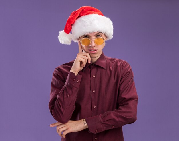 Réfléchi jeune homme blond portant bonnet de Noel et lunettes regardant la caméra faisant penser geste isolé sur fond violet