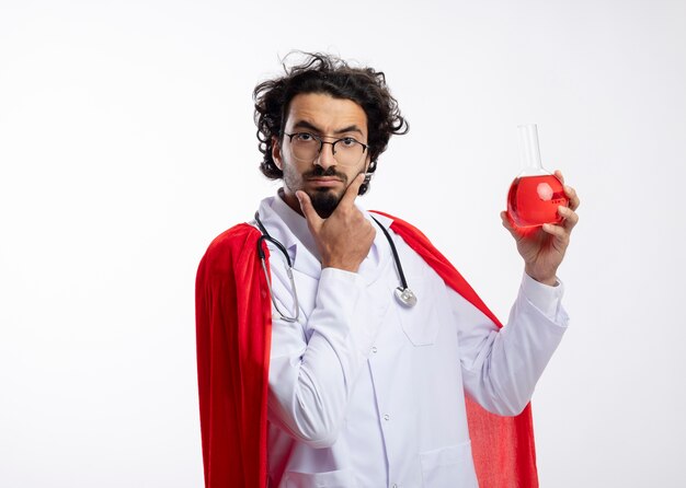 Réfléchi jeune homme caucasien à lunettes optiques portant l'uniforme du médecin avec manteau rouge et avec stéthoscope autour du cou met la main sur le menton et détient un liquide chimique rouge dans un flacon en verre