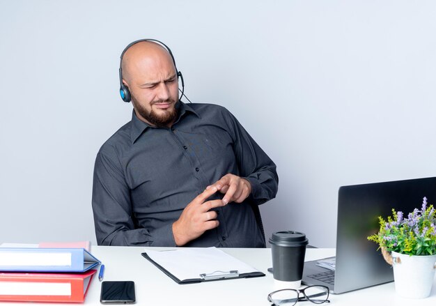 Réfléchi jeune homme de centre d'appels chauve portant un casque assis au bureau avec des outils de travail en gardant les doigts ensemble isolé sur un mur blanc