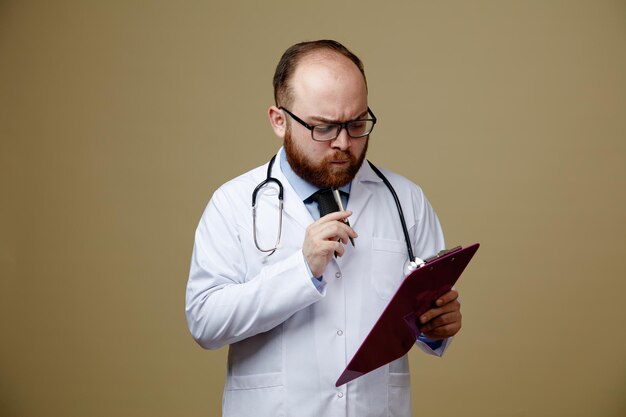 Réfléchi jeune homme médecin portant des lunettes blouse de laboratoire et stéthoscope autour du cou touchant le menton avec un stylo tenant et regardant le presse-papiers isolé sur fond vert olive