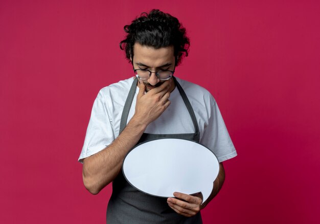 Réfléchi jeune homme de race blanche coiffeur portant des lunettes et bande de cheveux ondulés en uniforme tenant la bulle de chat mettant la main sur le menton regardant vers le bas isolé sur fond cramoisi avec espace de copie