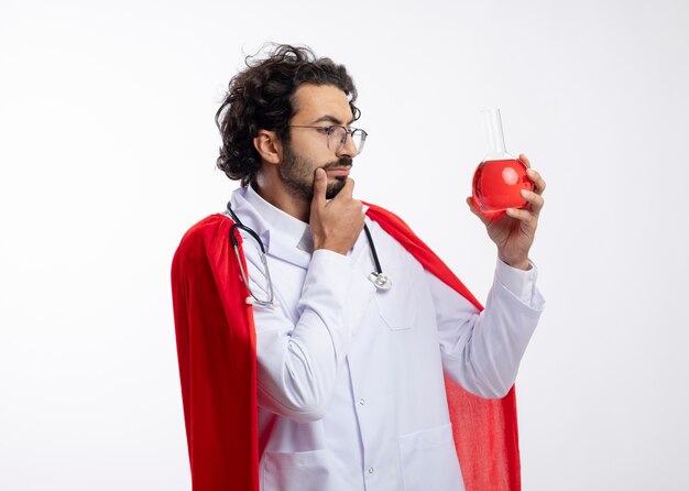 Réfléchi jeune homme de race blanche à lunettes optiques portant l'uniforme du médecin avec manteau rouge et avec stéthoscope autour du cou tient et regarde le liquide chimique rouge dans un flacon en verre