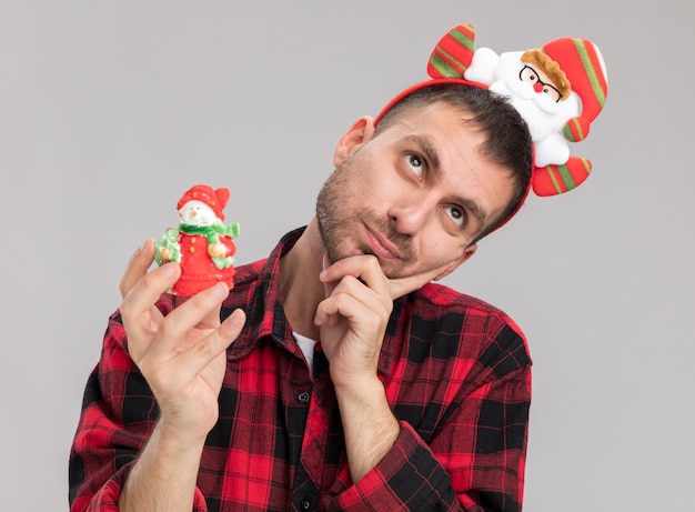 Réfléchi jeune homme de race blanche portant bandeau du père noël tenant ornement de Noël bonhomme de neige levant la main sur le menton isolé sur fond blanc