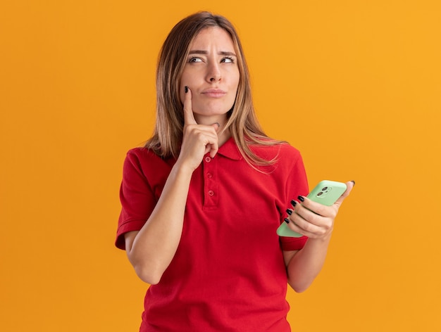 Photo gratuite réfléchi jeune jolie femme met le doigt sur le menton et tient le téléphone isolé sur un mur orange