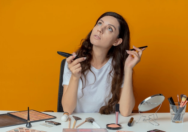 Réfléchi jeune jolie fille assise à la table de maquillage avec des outils de maquillage tenant eye-liner et mascara en levant isolé sur fond orange