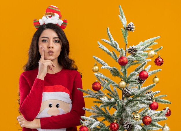 Réfléchi jeune jolie fille portant bandeau et pull du père noël debout près de l'arbre de Noël décoré regardant la caméra en gardant la main sur le menton pincer les lèvres isolé sur fond orange