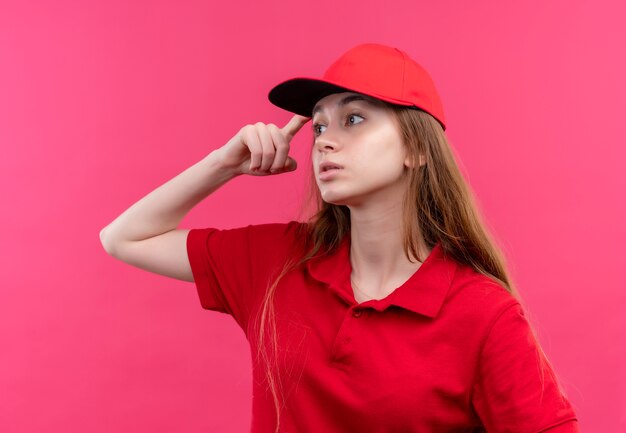 Réfléchi jeune livreuse en uniforme rouge mettant le doigt sur la tête en regardant le côté gauche sur un mur rose isolé