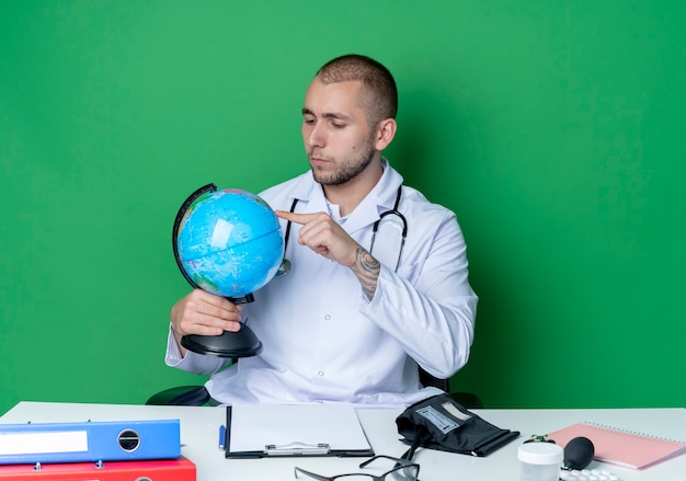 Réfléchi Jeune Médecin De Sexe Masculin Portant Une Robe Médicale Et Un Stéthoscope Assis Au Bureau Avec Des Outils De Travail Tenant Et Regardant Le Globe Et En Mettant Le Doigt Dessus Isolé Sur Un Mur Vert