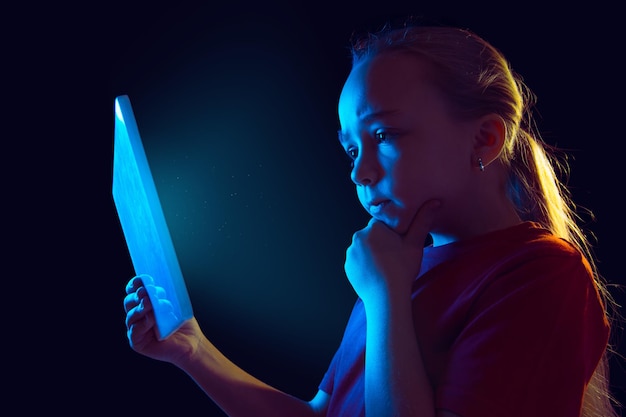 Réfléchi. Portrait de jeune fille caucasienne sur fond sombre de studio en néon. Beau modèle féminin à l'aide de tablette.