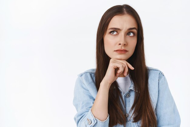 Réfléchi sérieux, déterminé et pensif, une fille des années 20 touchant le menton et fronçant les sourcils regarde dans le coin supérieur gauche en pensant face à un choix difficile debout sur fond blanc incertain et douteux