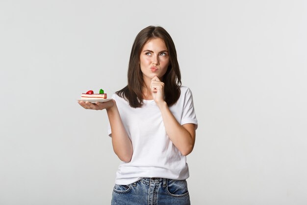 Réfléchi souriant jolie fille réfléchissant tout en tenant un morceau de gâteau, blanc.