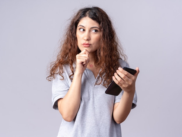 Réfléchie jeune jolie fille caucasienne tenant un téléphone portable en gardant la main sur le menton en regardant de côté