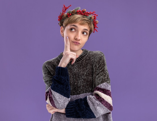 réfléchie jeune jolie fille portant une couronne de tête de noël touchant le visage regardant le côté isolé sur un mur violet avec espace de copie