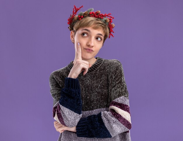 réfléchie jeune jolie fille portant une couronne de tête de noël touchant le visage regardant le côté isolé sur un mur violet avec espace de copie