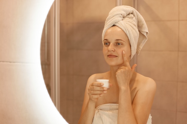 Reflet d'une jeune femme adulte dans un miroir appliquant une crème cosmétique sur le visage, mettant de l'argent nourrissant sur la peau de son visage dans la salle de bain, posant avec une serviette sur la tête.