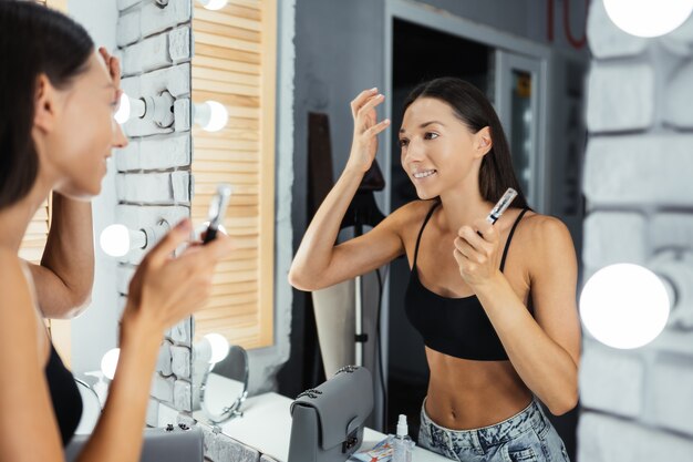 Réflexion de la belle jeune femme appliquant son maquillage, regardant dans un miroir