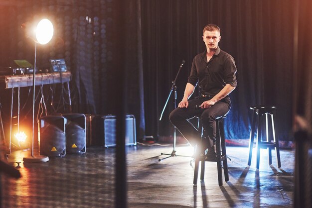 Regard magique. Beau jeune homme en vêtements noirs assis sur une chaise près de dans une pièce sombre avec de la lumière.