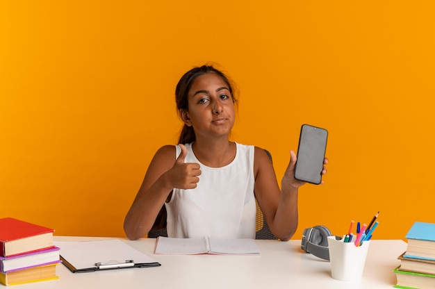 regardant la caméra jeune écolière assis au bureau avec des outils scolaires tenant le téléphone son pouce vers le haut