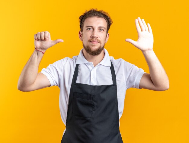 En regardant la caméra jeune homme barbier en uniforme montrant différents gestes isolés sur mur jaune