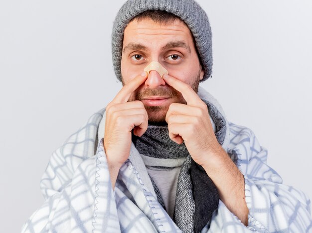 Regardant la caméra jeune homme malade portant un chapeau d'hiver et une écharpe enveloppée dans un plaid mettant du plâtre sur le nez isolé sur fond blanc