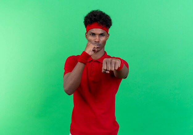 Regardant la caméra jeune homme sportif afro-américain portant bandeau et bracelet debout dans la pose de combat isolé sur fond vert