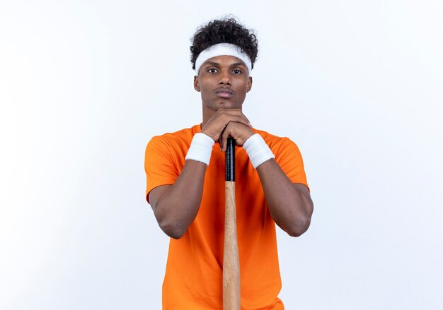 Regardant la caméra jeune homme sportif afro-américain portant bandeau et bracelet mettant le menton sur la chauve-souris isolé sur fond blanc