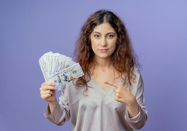 Photo gratuite regardant la caméra jeune jolie fille tenant et points à l'argent isolé sur fond bleu