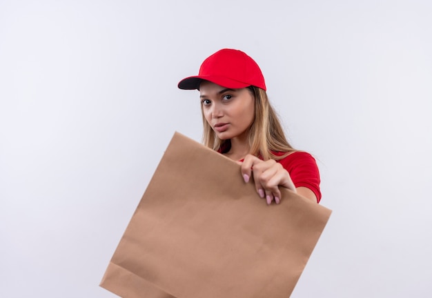 Photo gratuite regardant la caméra jeune livreuse portant un uniforme rouge et une casquette tenant un sac en papier isolé sur fond blanc